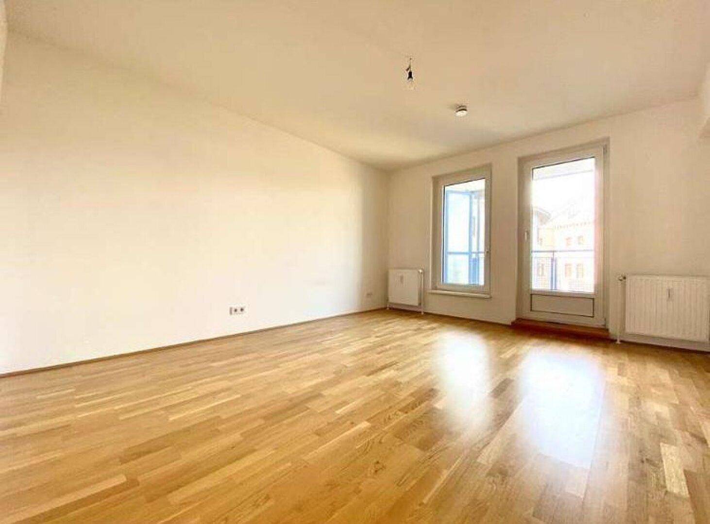 Heller, leerer Raum mit Parkettboden und großen Fenstern, die viel Tageslicht hereinlassen.