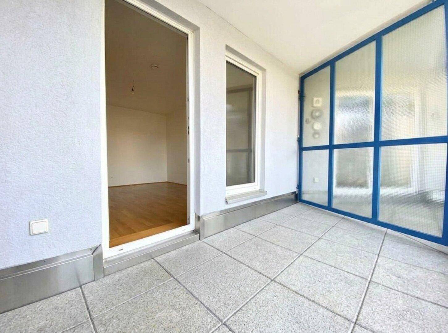 Heller Balkon mit Fliesenboden und blauer Glasabtrennung, zugänglich von einem Innenraum.