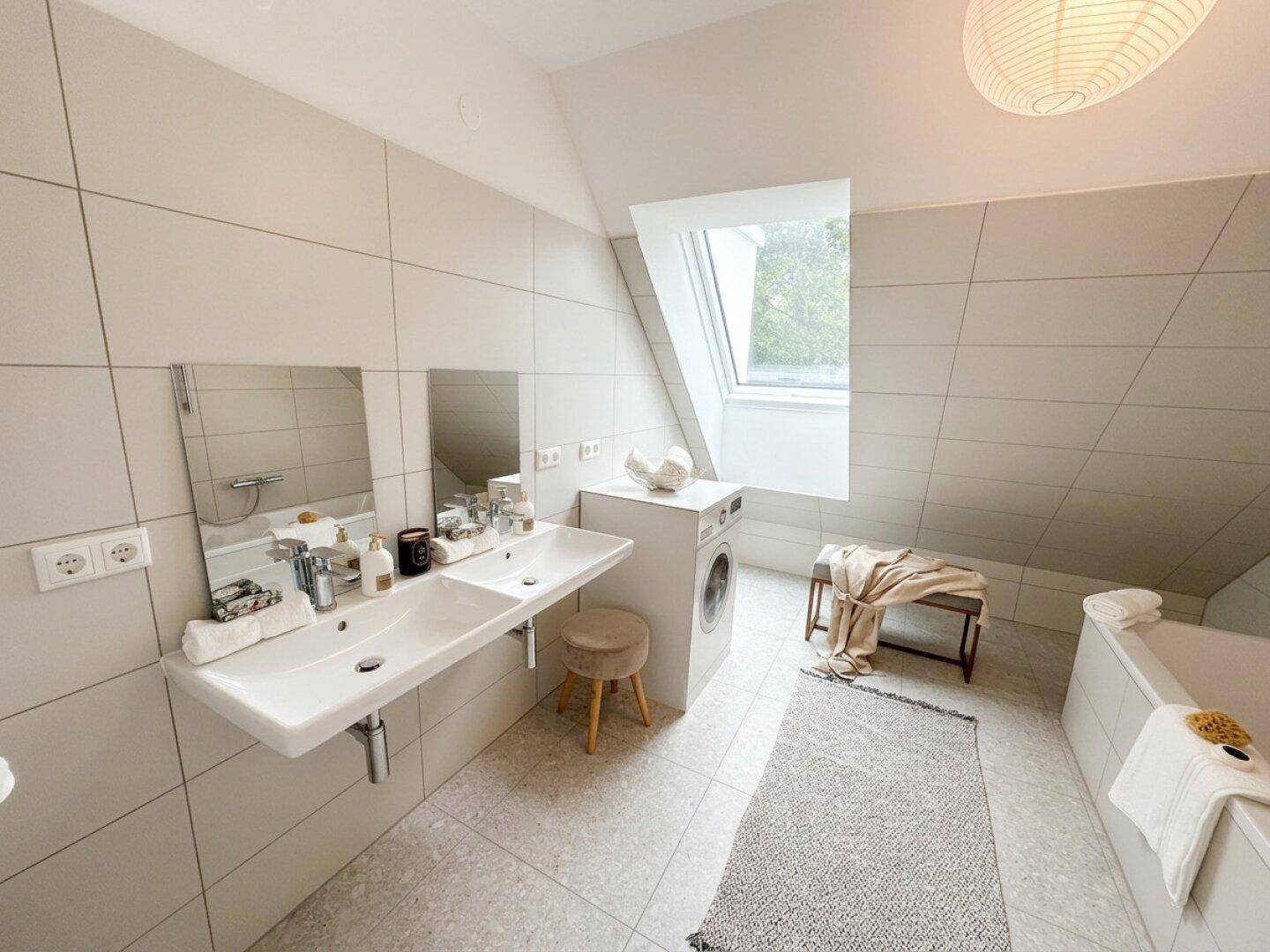 Geräumiges Badezimmer mit Doppelwaschbecken, Badewanne und Waschmaschine unter einem Dachfenster.