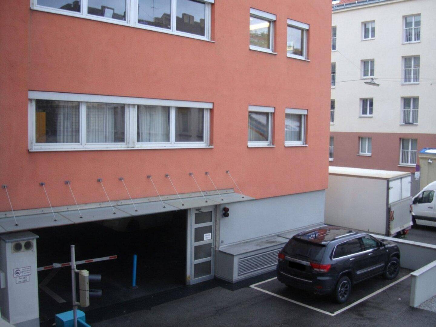 Außenansicht eines Gebäudes mit Tiefgarageneinfahrt, Außenparkplatz und Rollläden an den Fenstern.