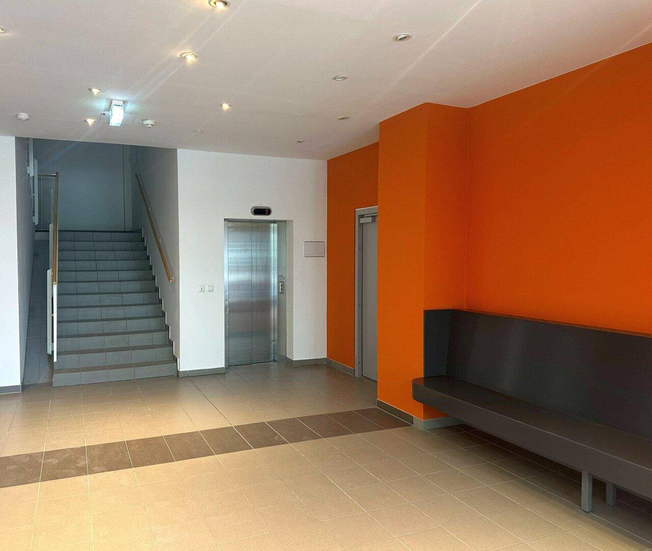 Empfangsbereich mit Aufzug, Treppe und Sitzgelegenheit, akzentuiert durch orangefarbene Wand.