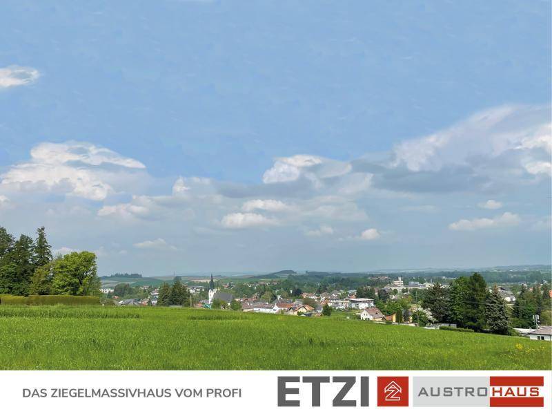 Panoramablick über grüne Felder auf eine ländliche Ortschaft mit Kirchturm und Hügeln.
