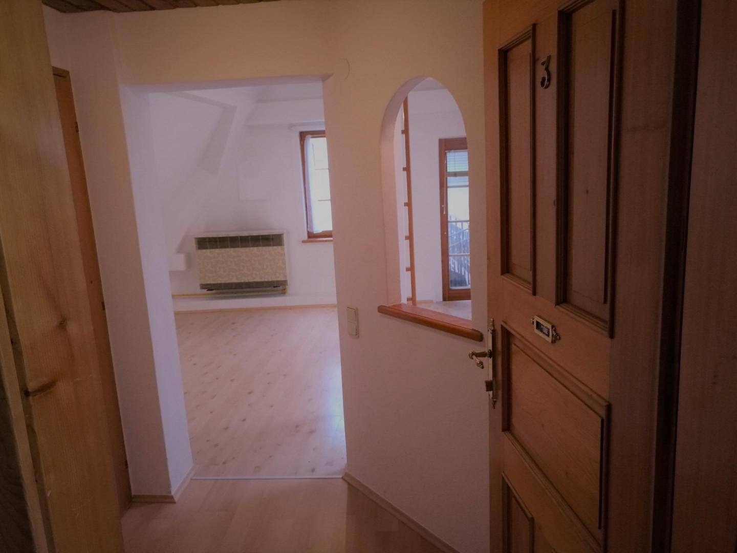 Flur mit Holztür und Blick in einen hellen Raum mit Heizkörper und Fenster unter Dachschräge.