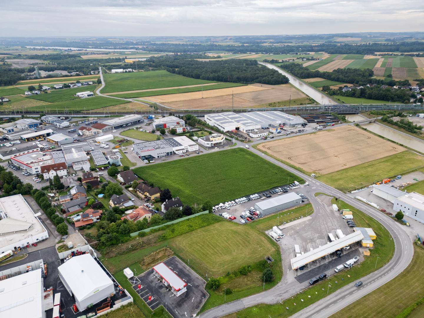 Weitläufige Luftaufnahme einer ländlichen Gegend mit Wohngebäuden, Gewerbeflächen, Feldern und einem Flusslauf unter bewölktem Himmel.