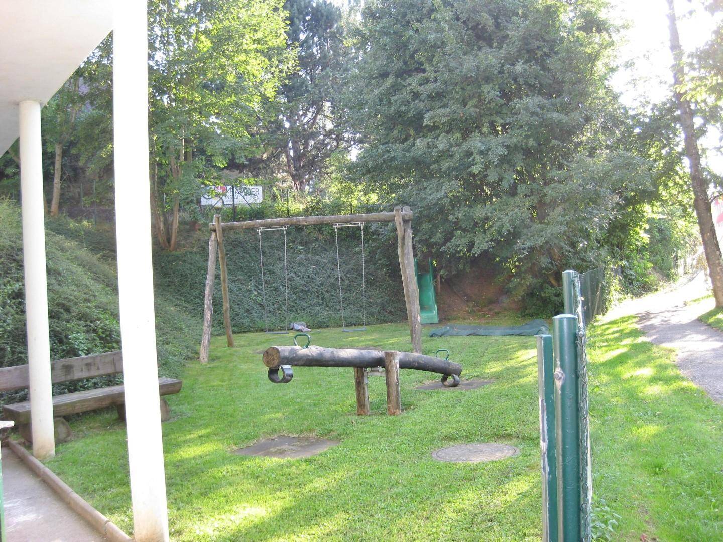 Spielplatz im Grünen mit Schaukel und Wippe, umgeben von Bäumen und Rasenflächen.