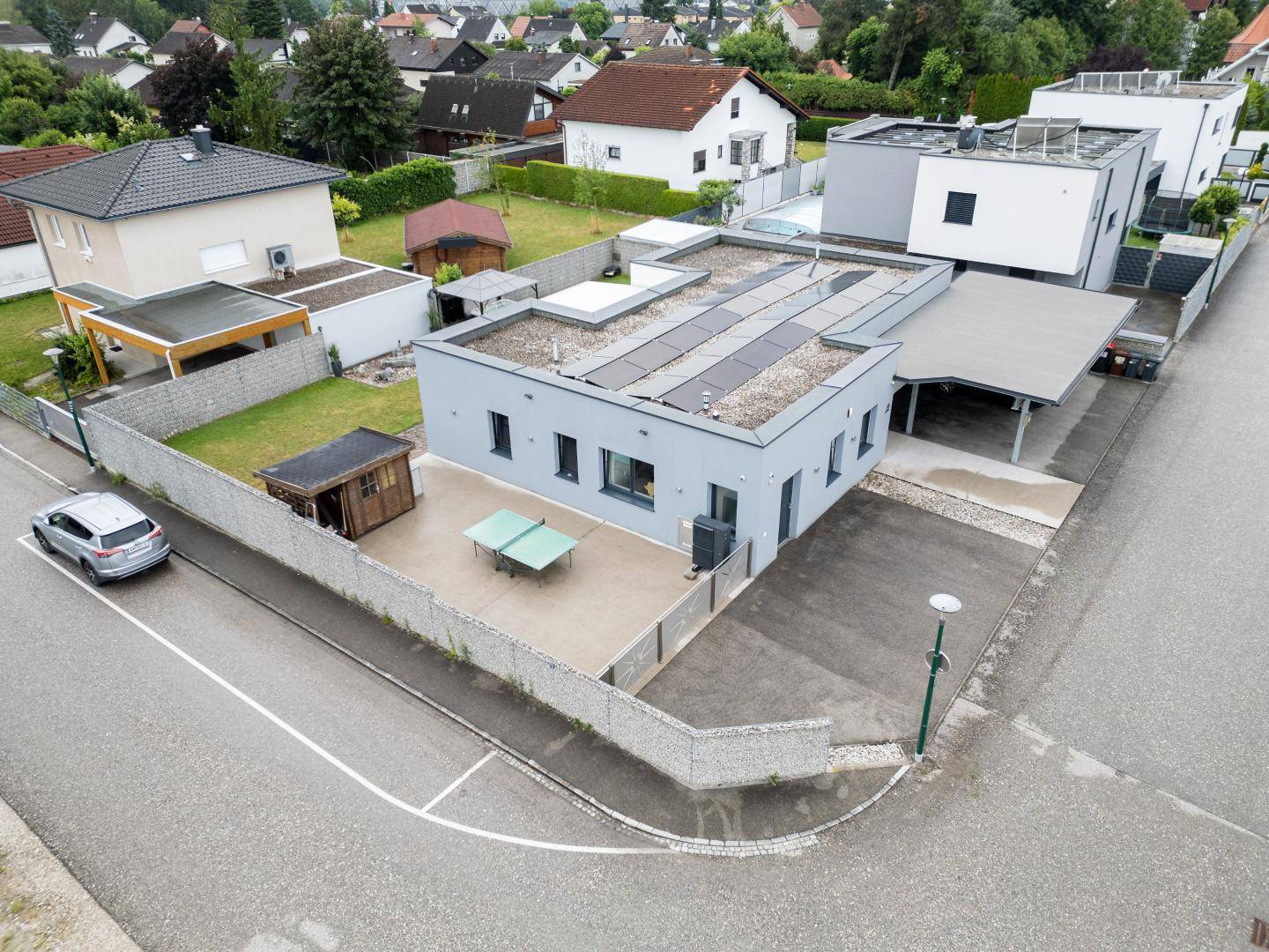 Luftaufnahme der Vorderseite des modernen Hauses mit Carport, gepflastertem Vorplatz und Tischtennisplatte.