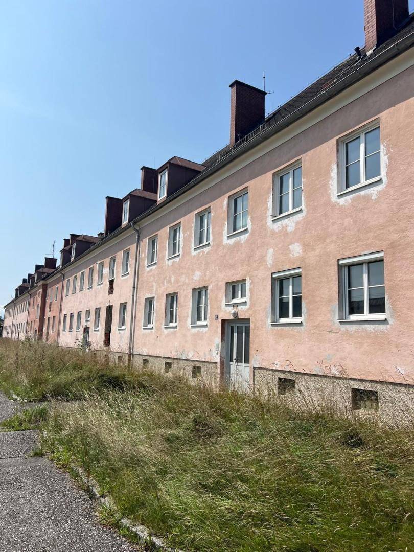 Nahaufnahme der verblassten rosa Fassade eines Wohngebäudes mit vielen Fenstern und hohem Gras im Vordergrund.