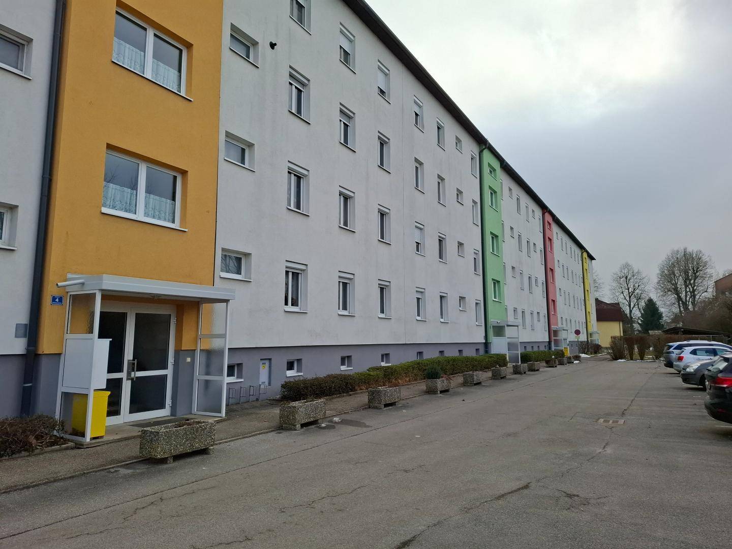 Lange Fassade eines Mehrfamilienhauses mit mehreren Eingängen und Parkplätzen.