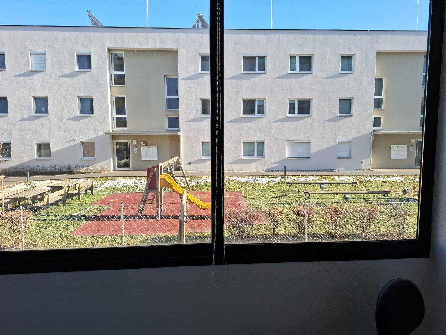 Blick aus dem Fenster auf einen Gemeinschaftsspielplatz mit Rutsche und Grünflächen.