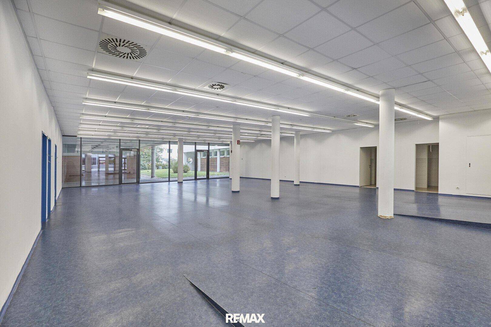 Heller Gewerberaum mit blauer Bodenfläche, weißen Säulen und Blick durch die Glasfront auf den Außenbereich.