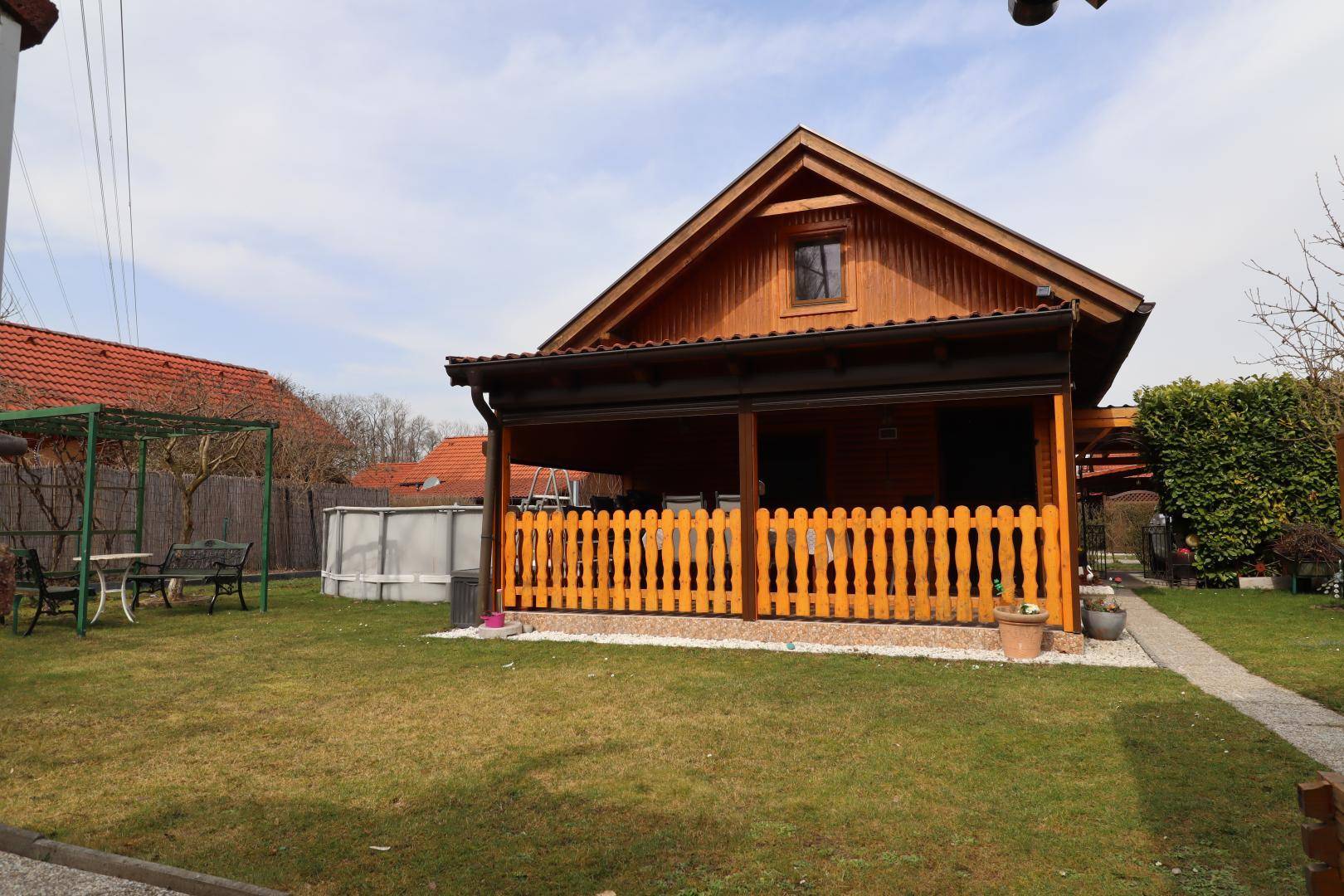Holzhaus mit überdachter Veranda, umgeben von einem grünen Garten und einem Pool.