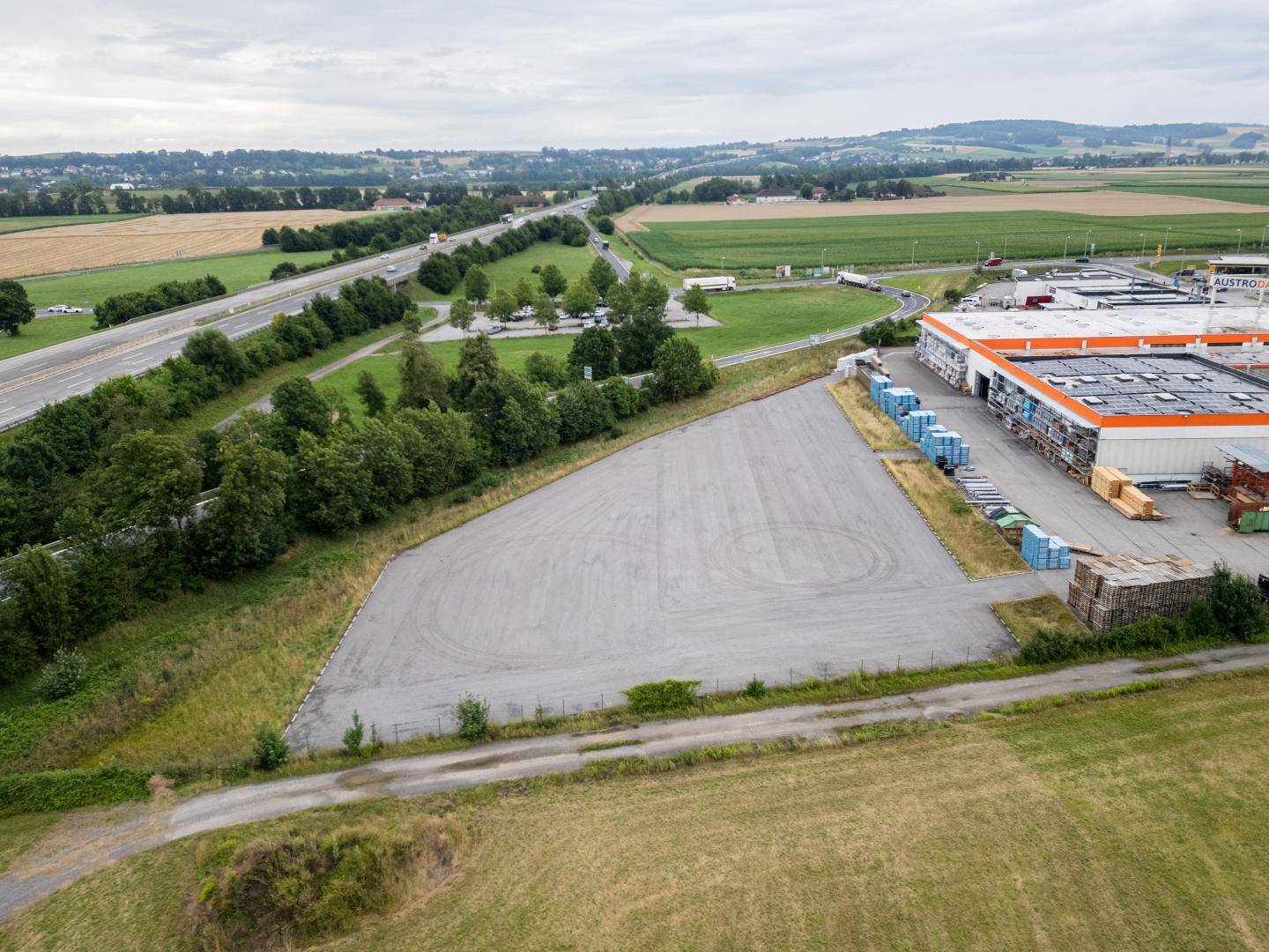 Luftaufnahme eines großen, dreieckigen Grundstücks neben einem Gewerbegebäude und einer Autobahn in ländlicher Umgebung.
