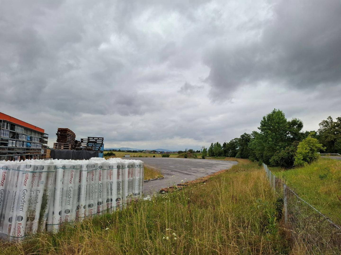 Eine gepflasterte Fläche mit Baumaterialien und Paletten im Vordergrund, angrenzend an ein Gewerbegebäude und grüne Landschaft.