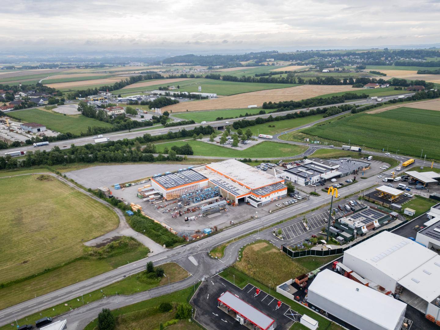 Weitläufige Luftaufnahme eines Gewerbegebiets mit mehreren Gebäuden, Straßen und einer Autobahn, umgeben von Feldern.