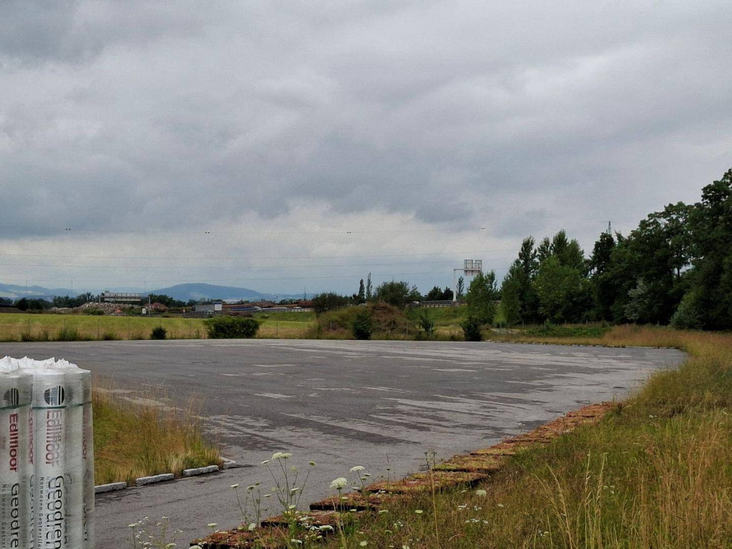 Eine große, gepflasterte Fläche mit Baumaterialien im Vordergrund, umgeben von Gras und Bäumen unter bewölktem Himmel.