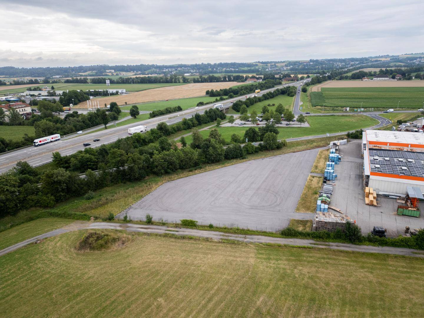 Luftaufnahme eines großen, dreieckigen Grundstücks neben einem Gewerbegebäude und einer Autobahn in ländlicher Umgebung.