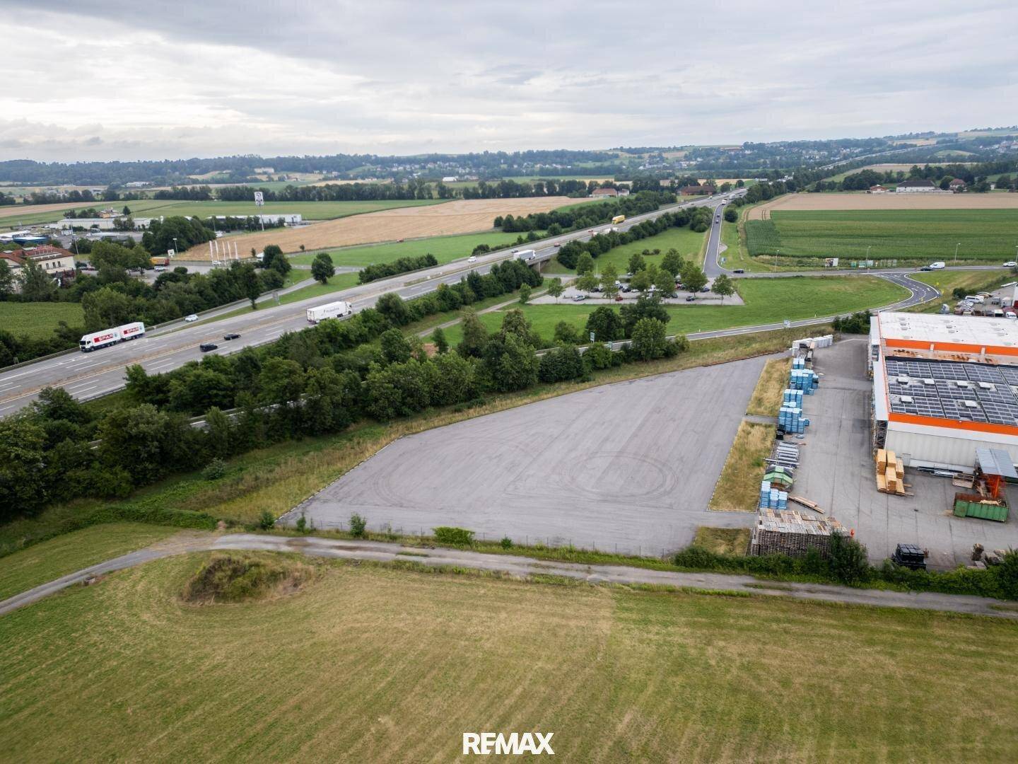 Luftaufnahme einer großen asphaltierten Fläche neben einem Gewerbegebäude und einer Autobahn, umgeben von Feldern.