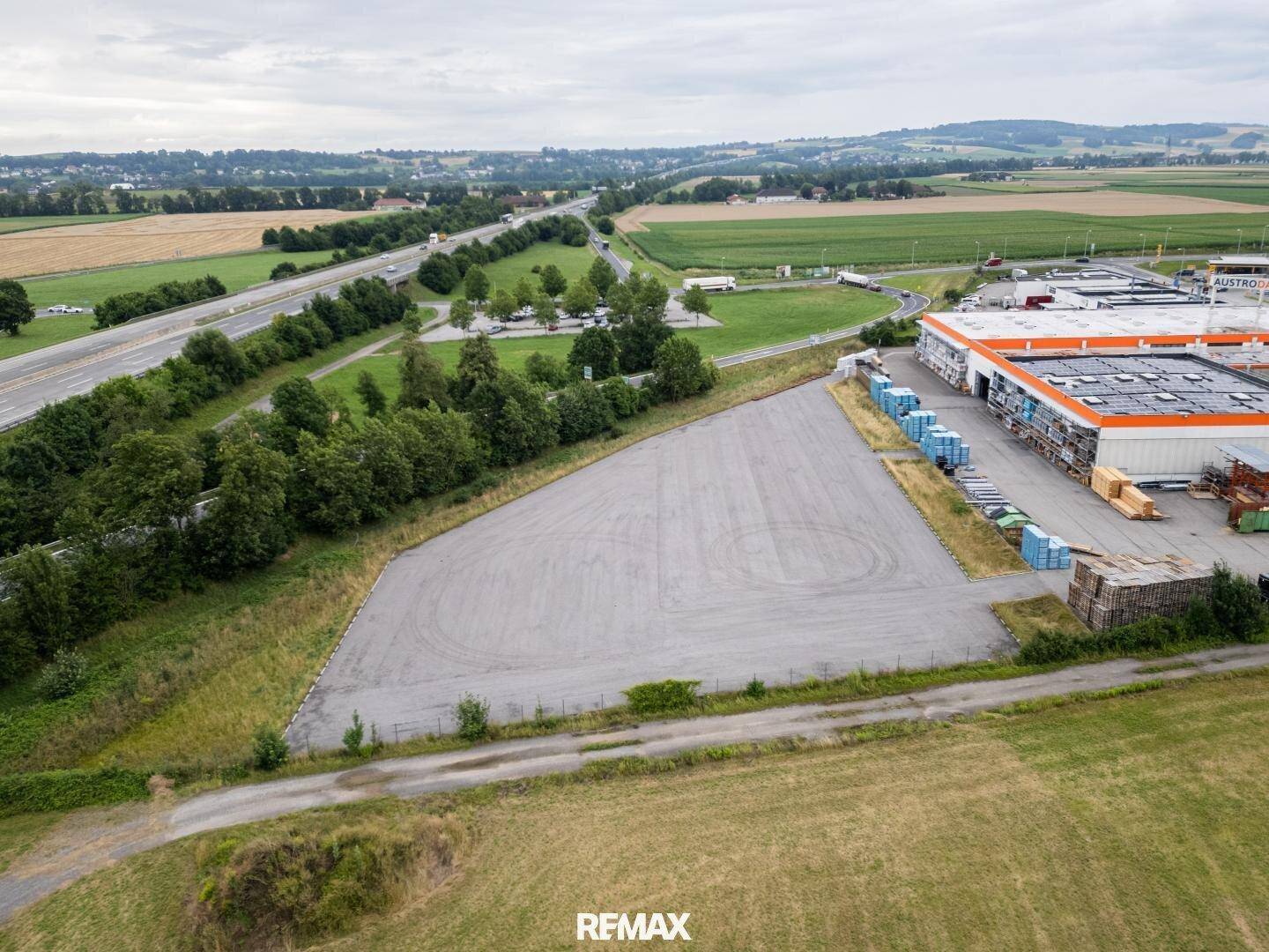 Luftaufnahme einer großen asphaltierten Fläche neben einem Gewerbegebäude und einer Autobahn, umgeben von Feldern.