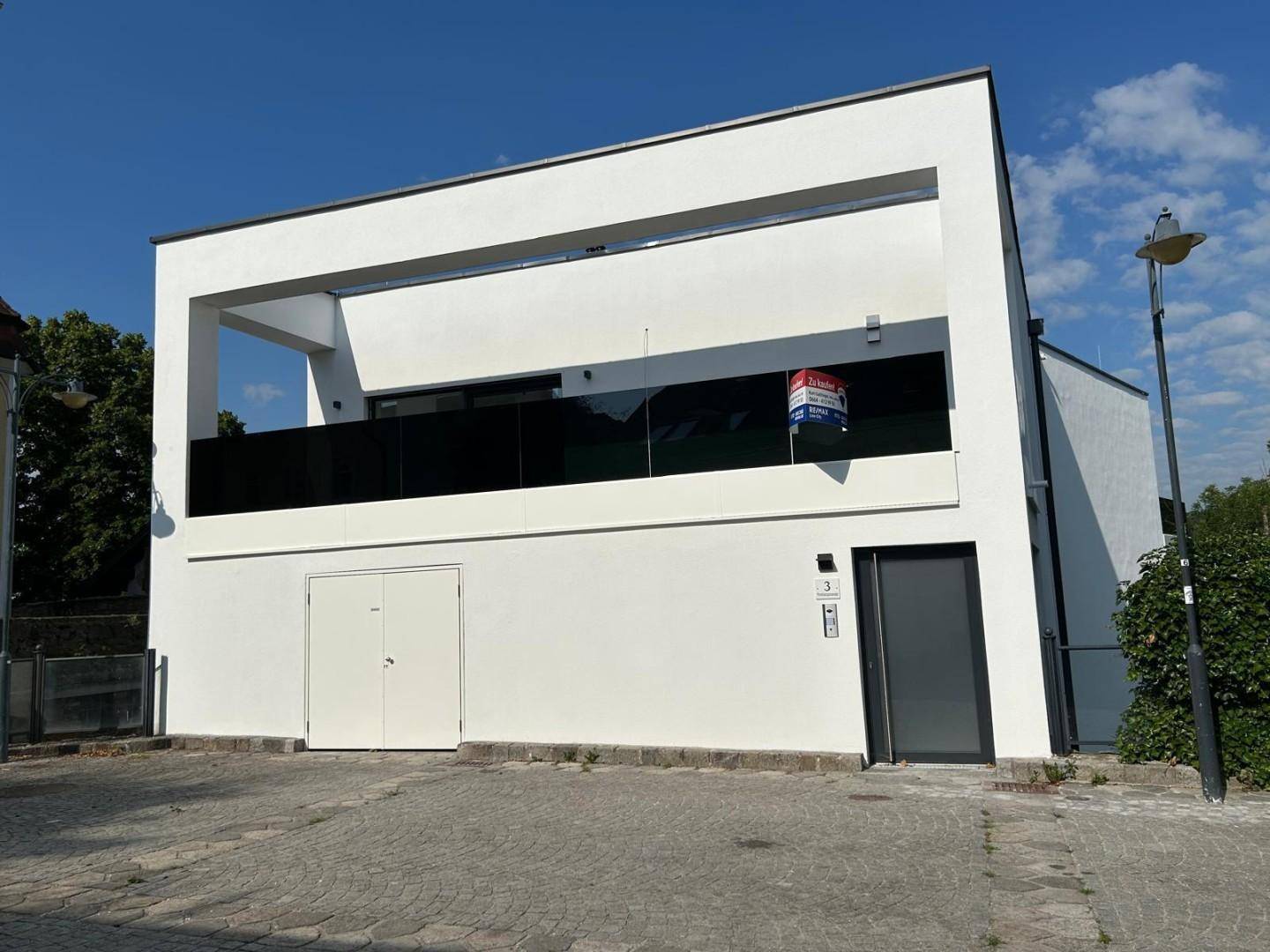Moderne weiße Fassade eines Neubaus mit Balkon und einem 'Zu verkaufen'-Schild.
