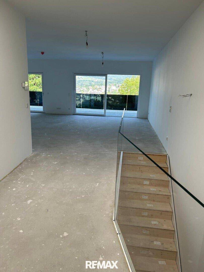 Heller Flur mit Betonboden, Holztreppe und Glasgeländer, mit Blick ins Grüne.