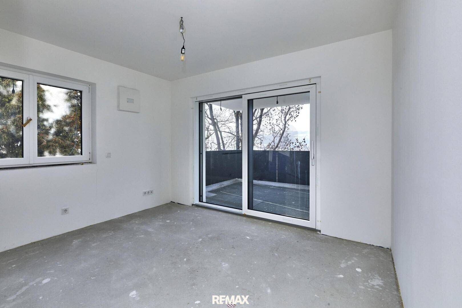 Helles, unfertiges Zimmer mit Betonboden, Fenster und Zugang zu einem kleinen Balkon.