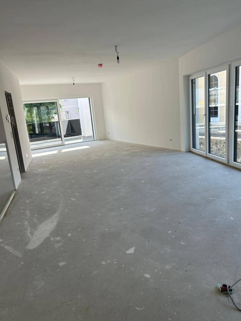 Großer, heller Wohnbereich im Rohbau mit vielen Fenstern, die viel Tageslicht hereinlassen.