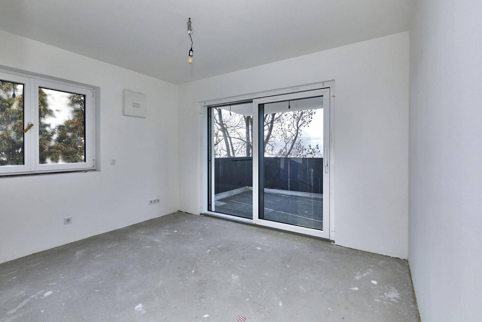 Helles Schlafzimmer im Rohbau mit Fenster und Zugang zu einem Balkon, bereit für den Ausbau.