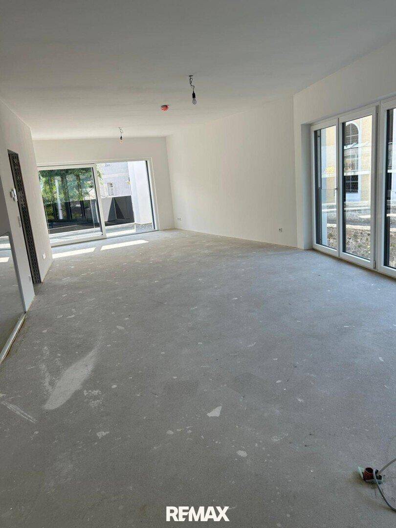 Großer, unfertiger Wohnbereich mit Betonboden und vielen Fenstern für natürliches Licht.