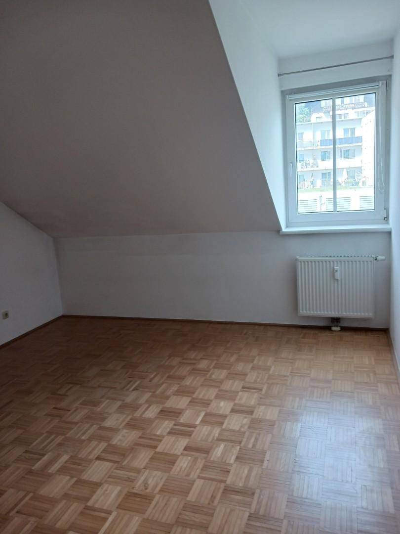 Heller Raum mit Parkettboden, Dachschräge und Fenster, das einen Blick auf die umliegenden Gebäude bietet.