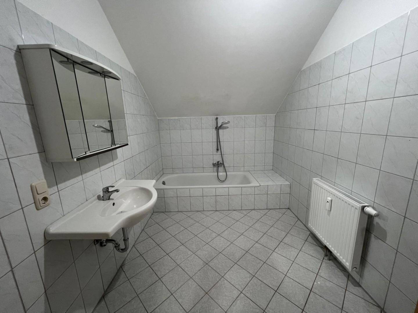 Badezimmer mit hellgrauen Fliesen, Badewanne mit Duschfunktion, Waschbecken und Spiegelschrank unter Dachschräge.
