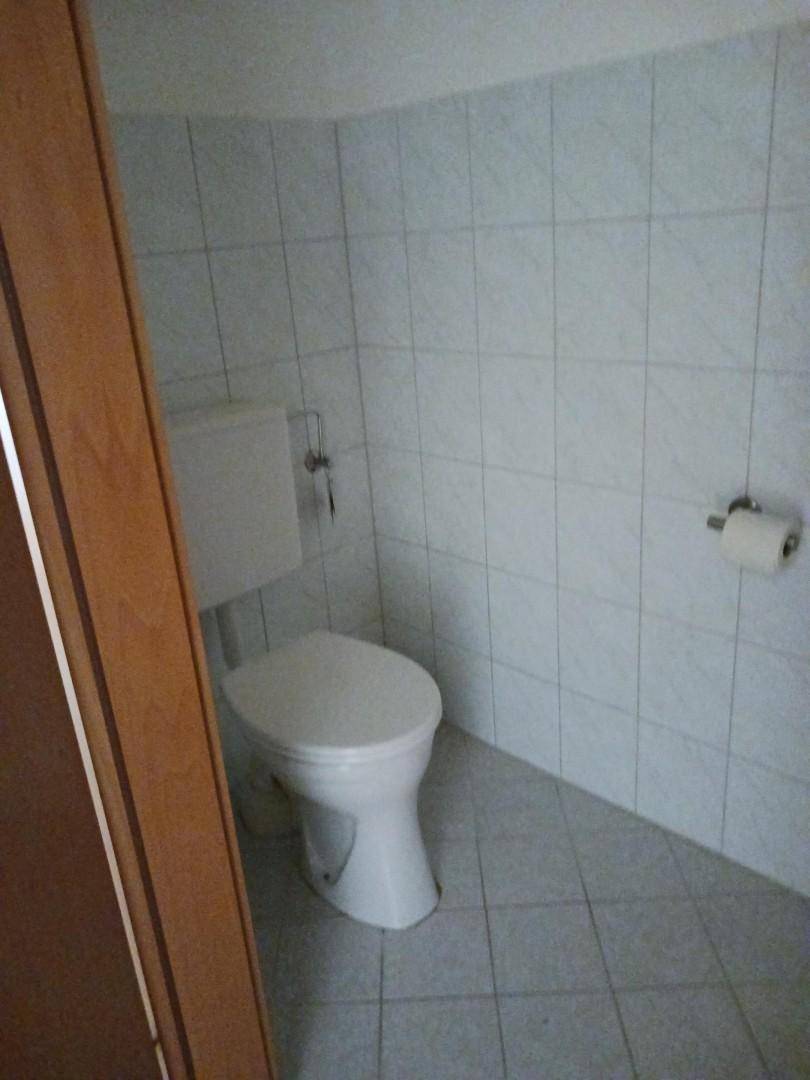Kleines Gäste-WC mit hellgrauen Fliesen an Wänden und Boden, ausgestattet mit einer einfachen Toilette.
