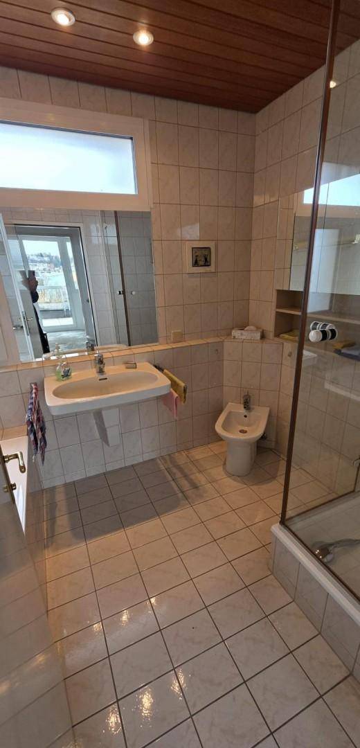 Badezimmer mit hellen Fliesen, Einzelwaschbecken, Bidet und einer begehbaren Dusche mit Glaswand.