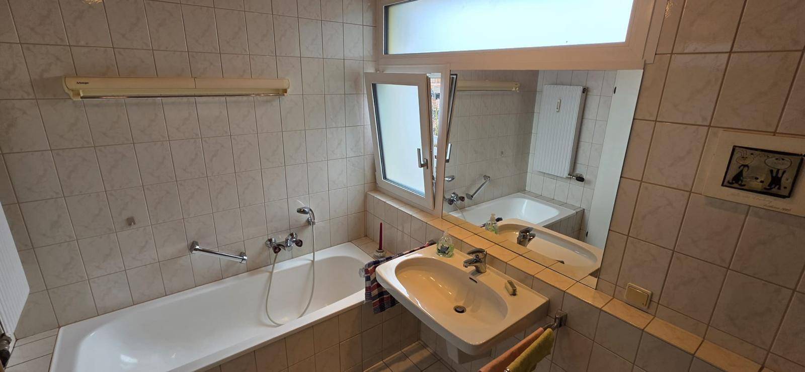 Badezimmer mit Badewanne, Einzelwaschbecken und Fenster, ausgestattet mit hellen Fliesen und Holzdecke.