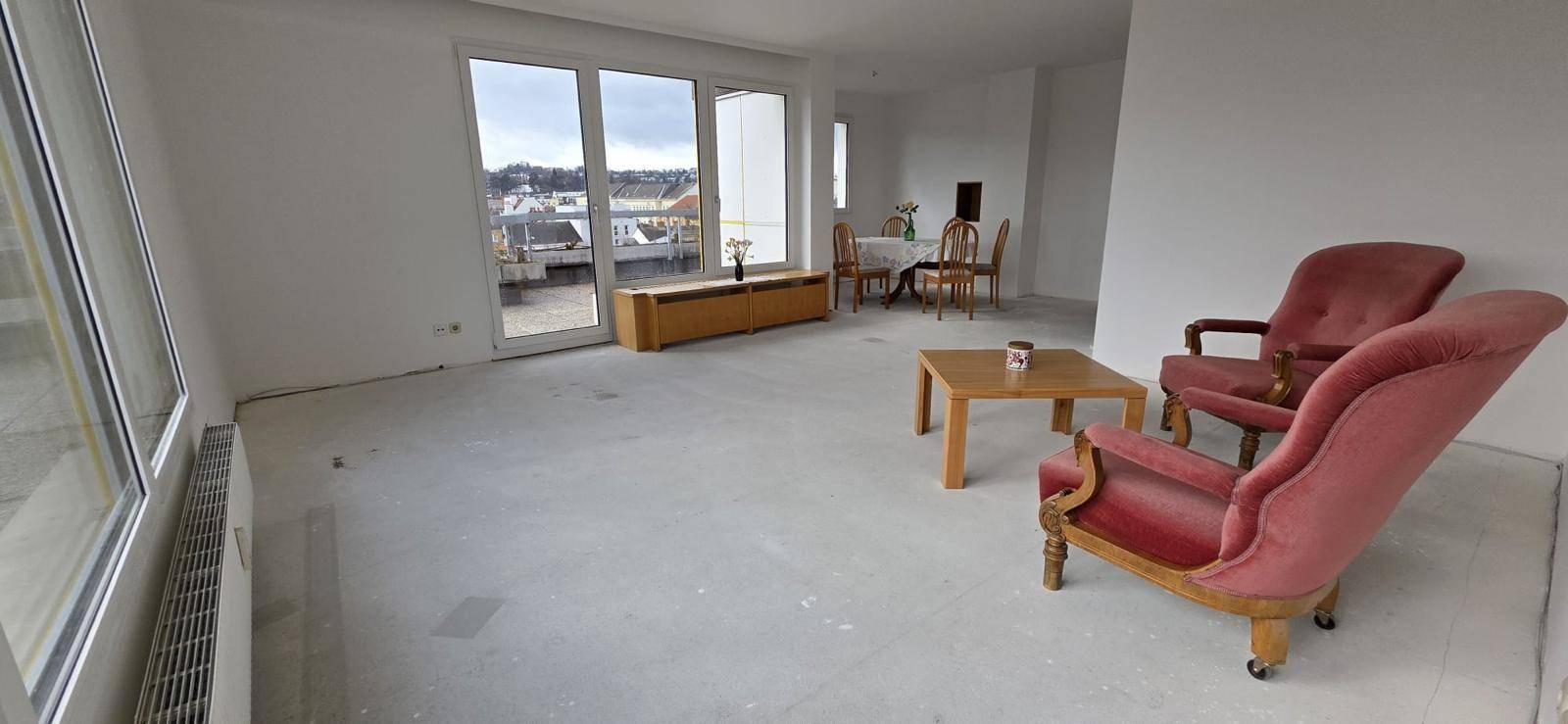 Geräumiger Wohnbereich mit großen Fenstern, Betonboden und Vintage-Sesseln, bereit zur individuellen Einrichtung.