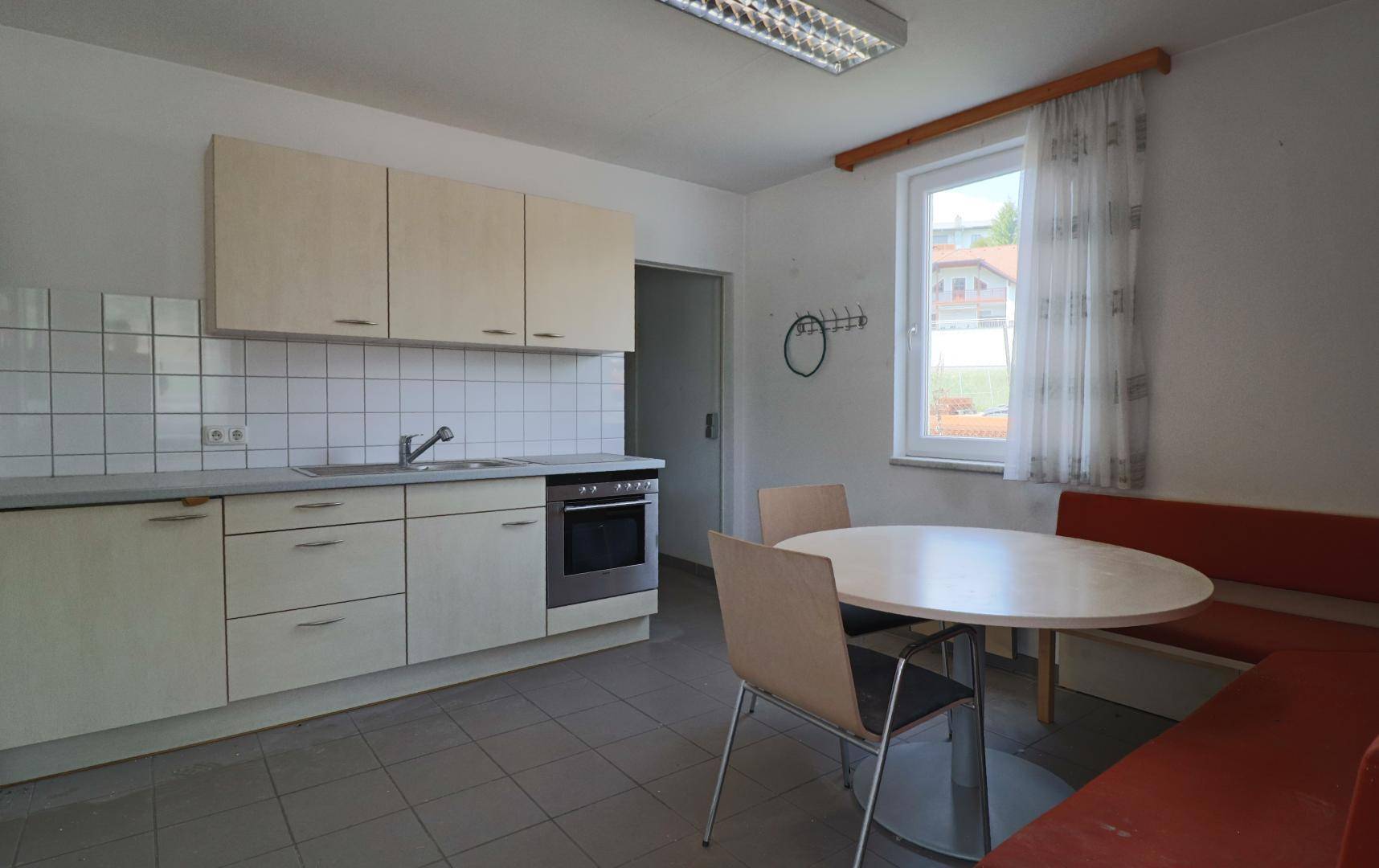 Funktionale Küche mit hellen Einbauschränken, einem Esstisch mit Bank und Fliesenboden, gut beleuchtet durch ein Fenster.