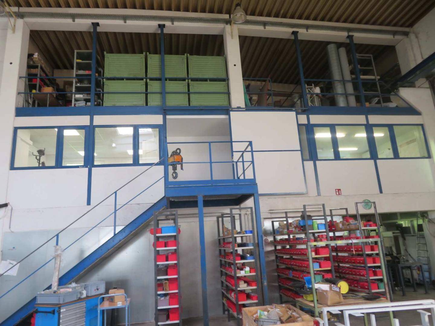 Mehrstöckige interne Struktur mit Bürobereichen, Lagerregalen und einer blauen Metalltreppe in einer Halle.