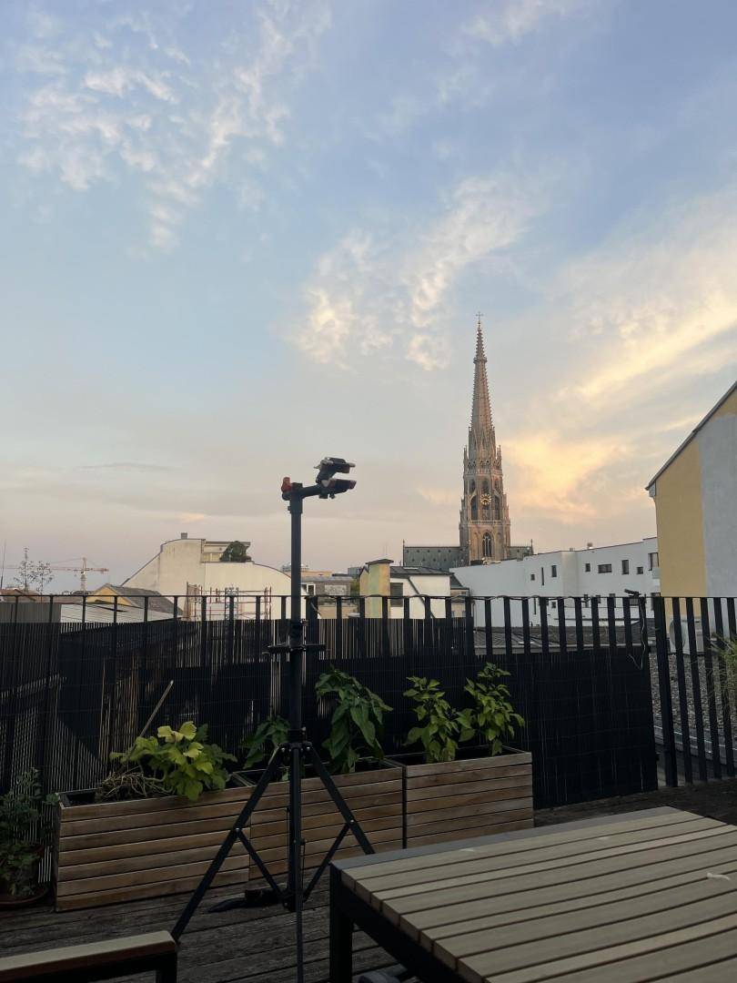 Geräumige Dachterrasse mit Holztisch, Pflanzen und weitem Blick auf eine historische Kirchturmspitze bei Sonnenuntergang.