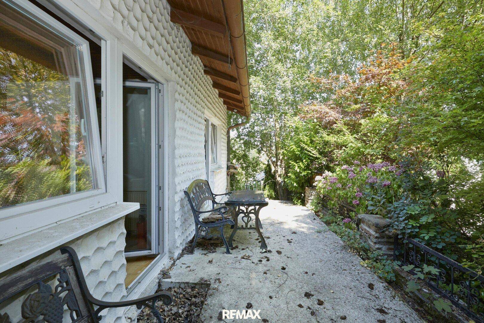 Einfache Terrasse mit Gartenmöbeln neben dem Haus, umgeben von Bäumen und Sträuchern.