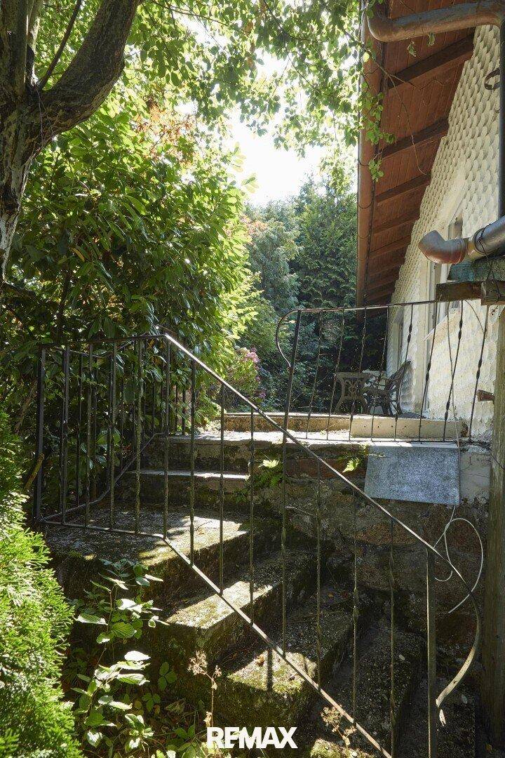 Steintreppe mit schmiedeeisernem Geländer führt zum Hauseingang, umgeben von üppiger Vegetation.