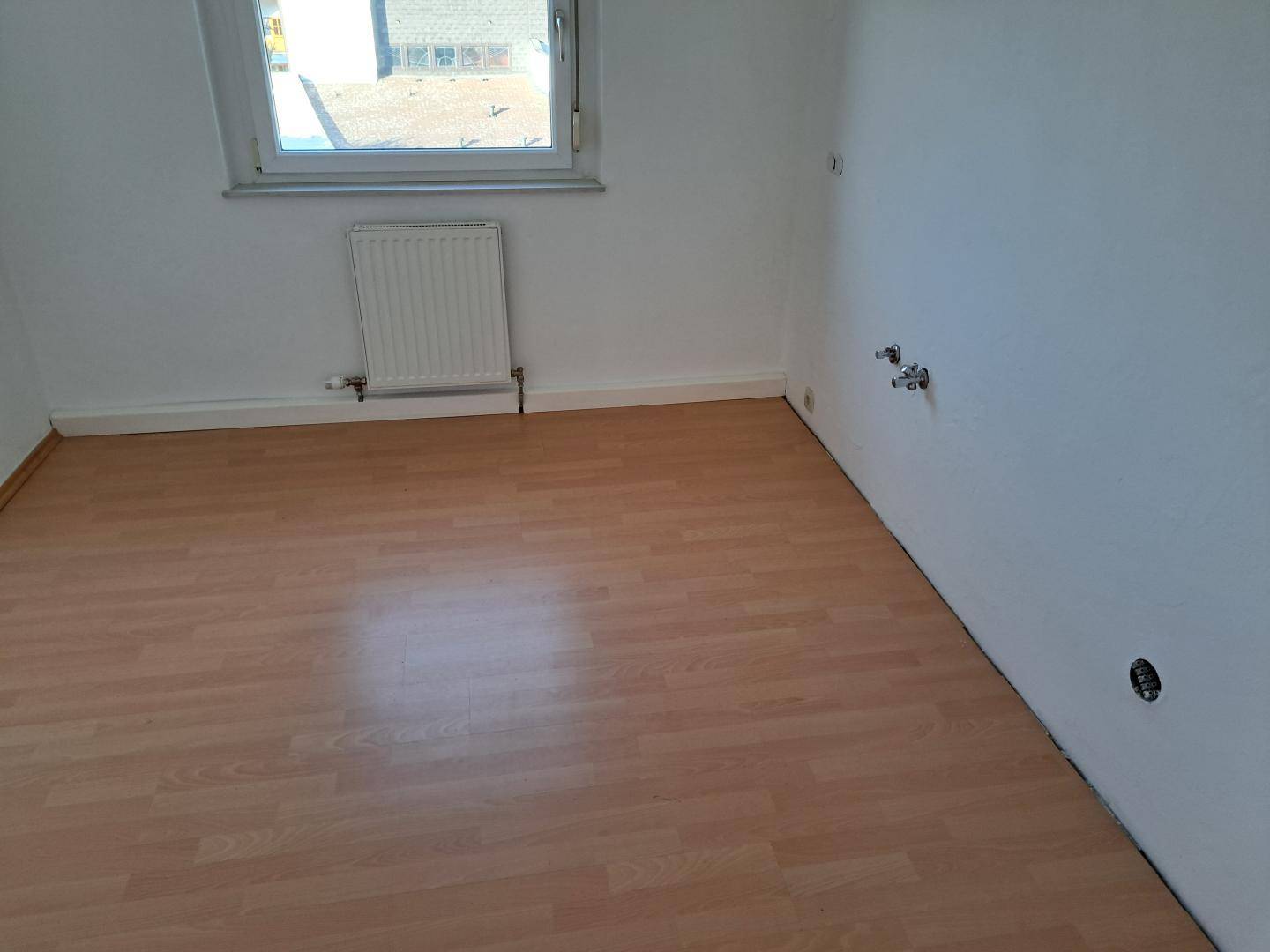Leeres Zimmer mit Laminatboden, Fenster, Heizkörper und vorbereiteten Wasseranschlüssen an der Wand.