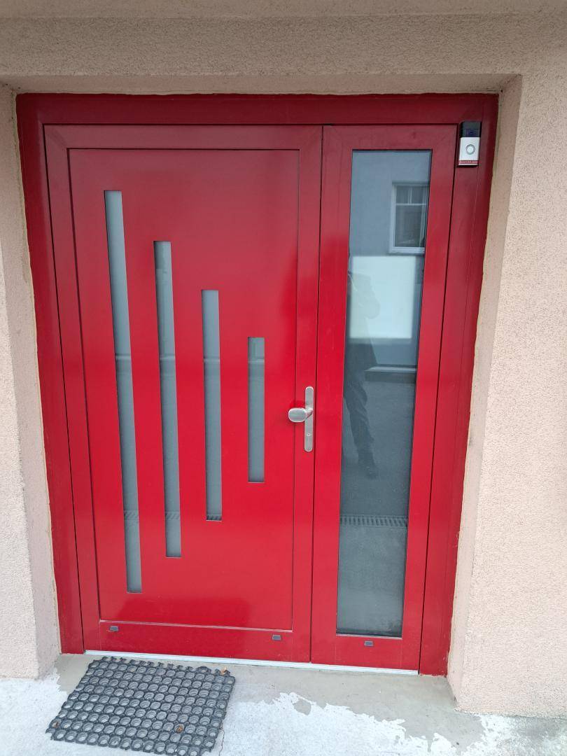 Moderne rote Eingangstür mit vertikalen Glaselementen und einem kleinen Fußabtreter davor.