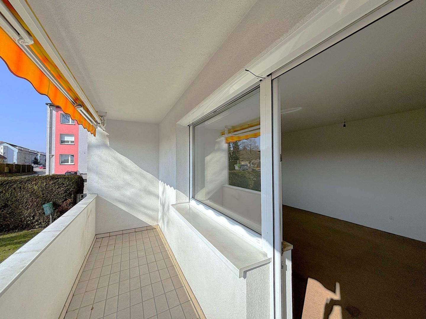 Sonniger Balkon mit Fliesenboden, weißem Geländer und orangefarbener Markise, mit Blick auf die Umgebung.