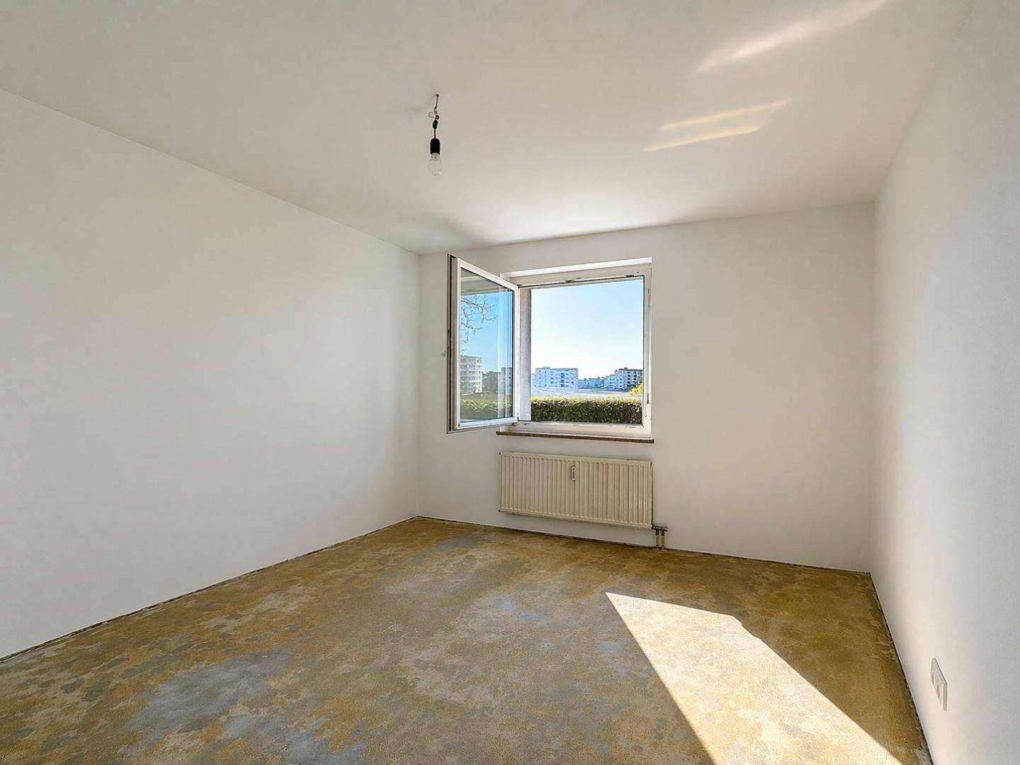 Helles, leeres Schlafzimmer mit Rohboden, weißen Wänden und einem Fenster mit Blick ins Grüne.