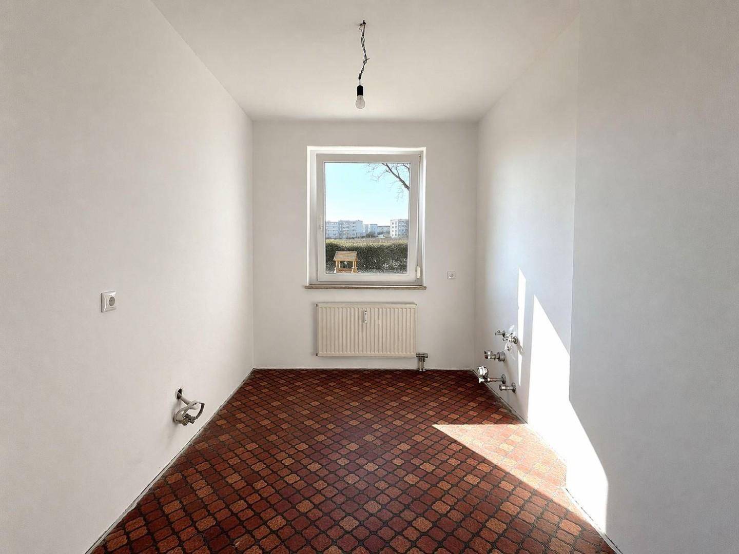 Leere Küche mit braunen Fliesen, weißen Wänden, Fenster und Anschlüssen für Küchengeräte.