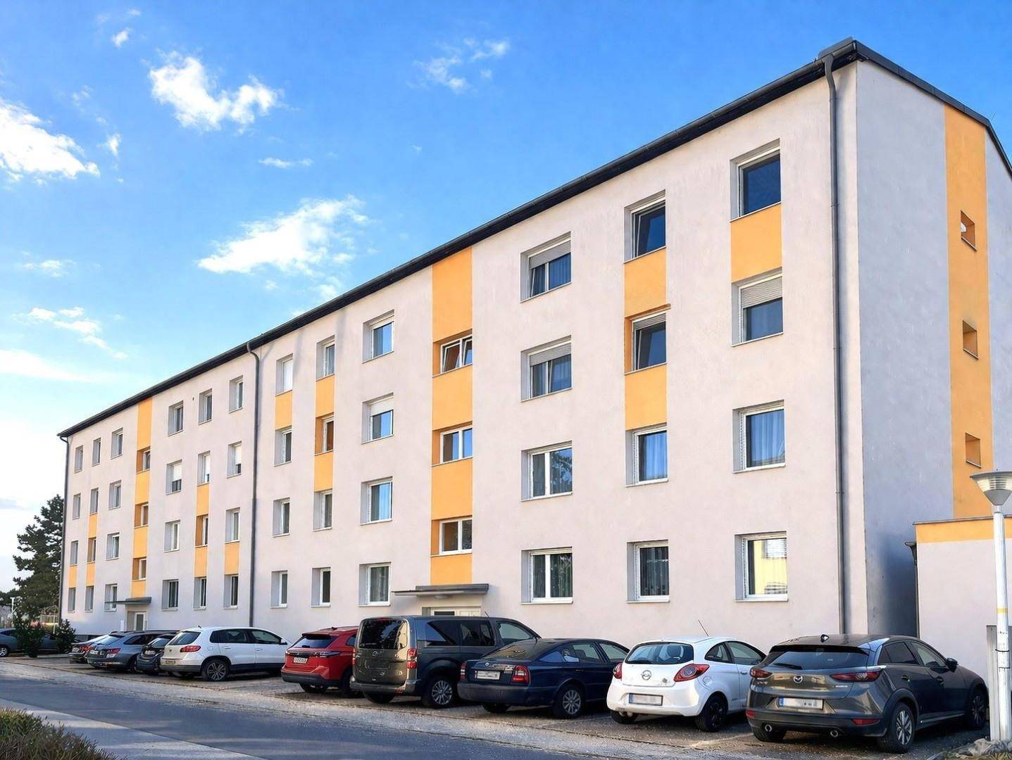 Mehrstöckiges Wohngebäude mit grauer und orangefarbener Fassade und Parkplätzen davor unter blauem Himmel.
