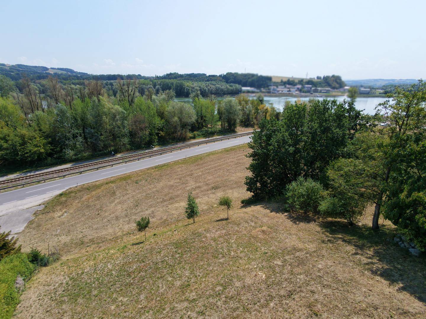 Blick über trockenes Grasland auf den Fluss, umgeben von Bäumen, einer Straße und Bahnlinie.