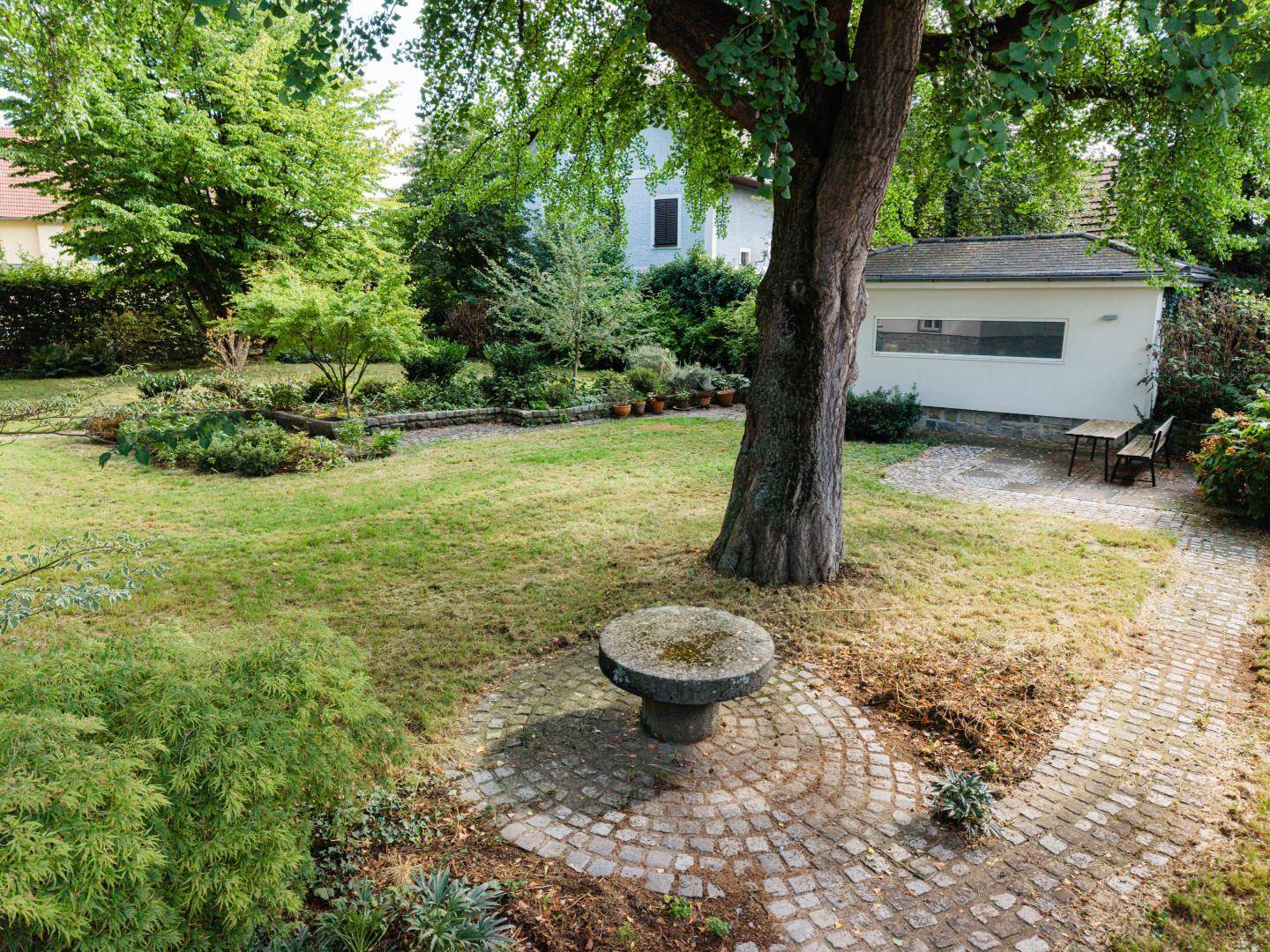 Weitläufiger Garten mit Rasen, altem Baumbestand, einem Steintisch und einem Gartenhaus.