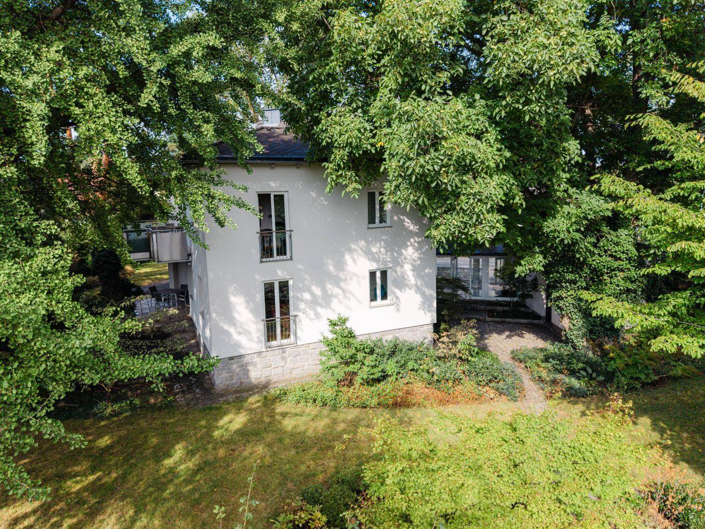 Rückansicht des weißen Hauses, umgeben von üppigem Grün und hohen Bäumen.