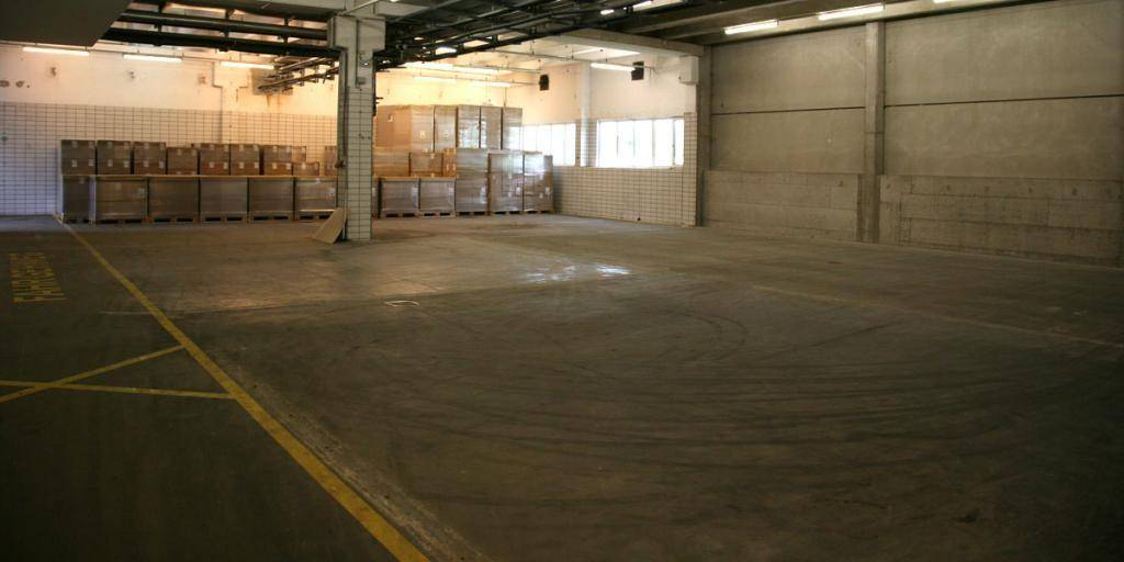 Große, leere Lagerhalle mit Betonboden, hohen Decken und gestapelten Kisten im Hintergrund.