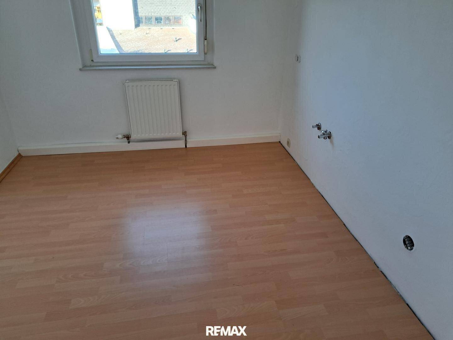 Leerer Raum mit Laminatboden und vorbereiteten Anschlüssen für eine Küche.